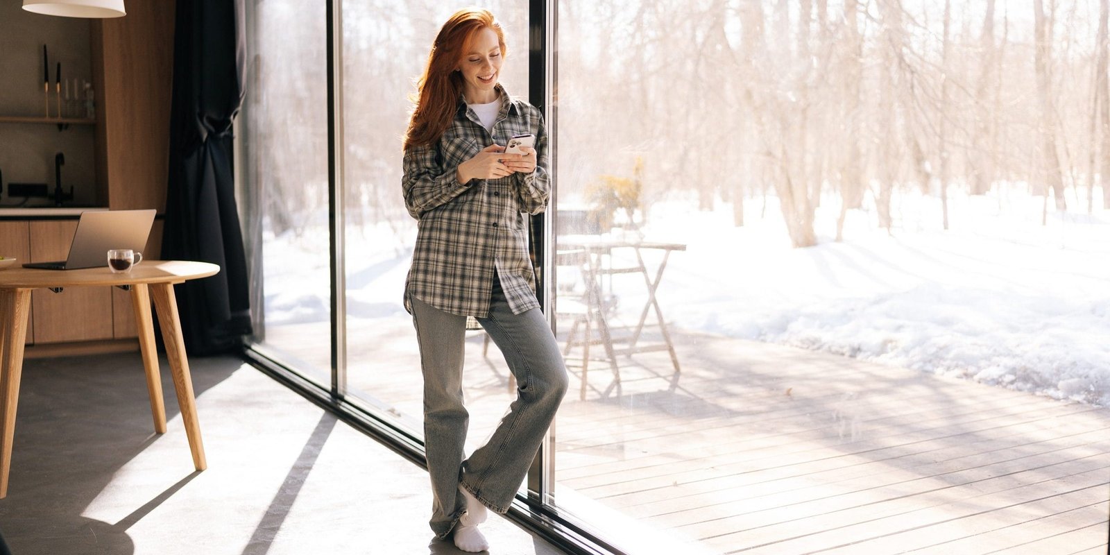 Full length portrait of young woman in casual clothes using smartphone looking to screen standing by window at home on sunny day. Happy redhead female enjoying using mobile